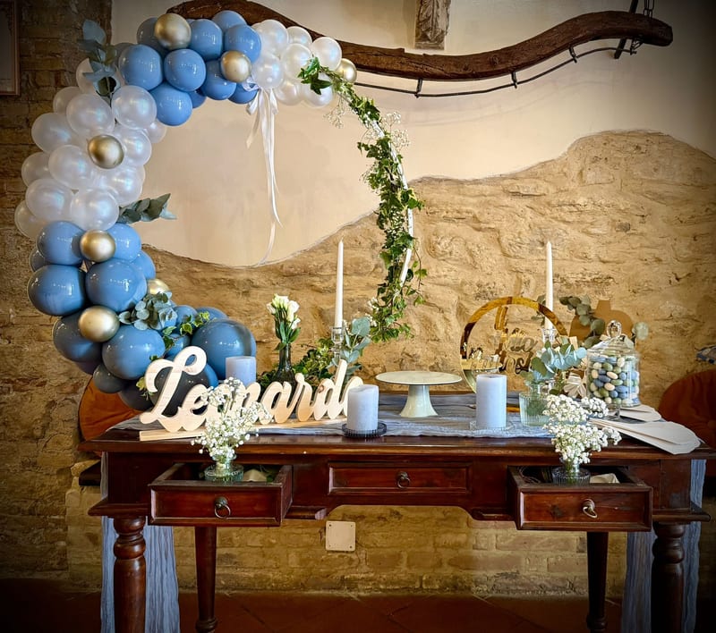 Tavolo dolci per battesimo con arco floreale, palloncini azzurri e scritta in legno